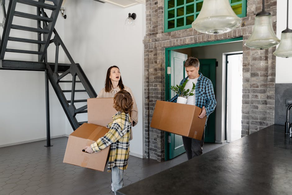 A young woman with curly black hair and a bright smile is sitting on a wooden floor inside a room, surrounded by large cardboard boxes used for packing belongings during a home relocation. She is wear